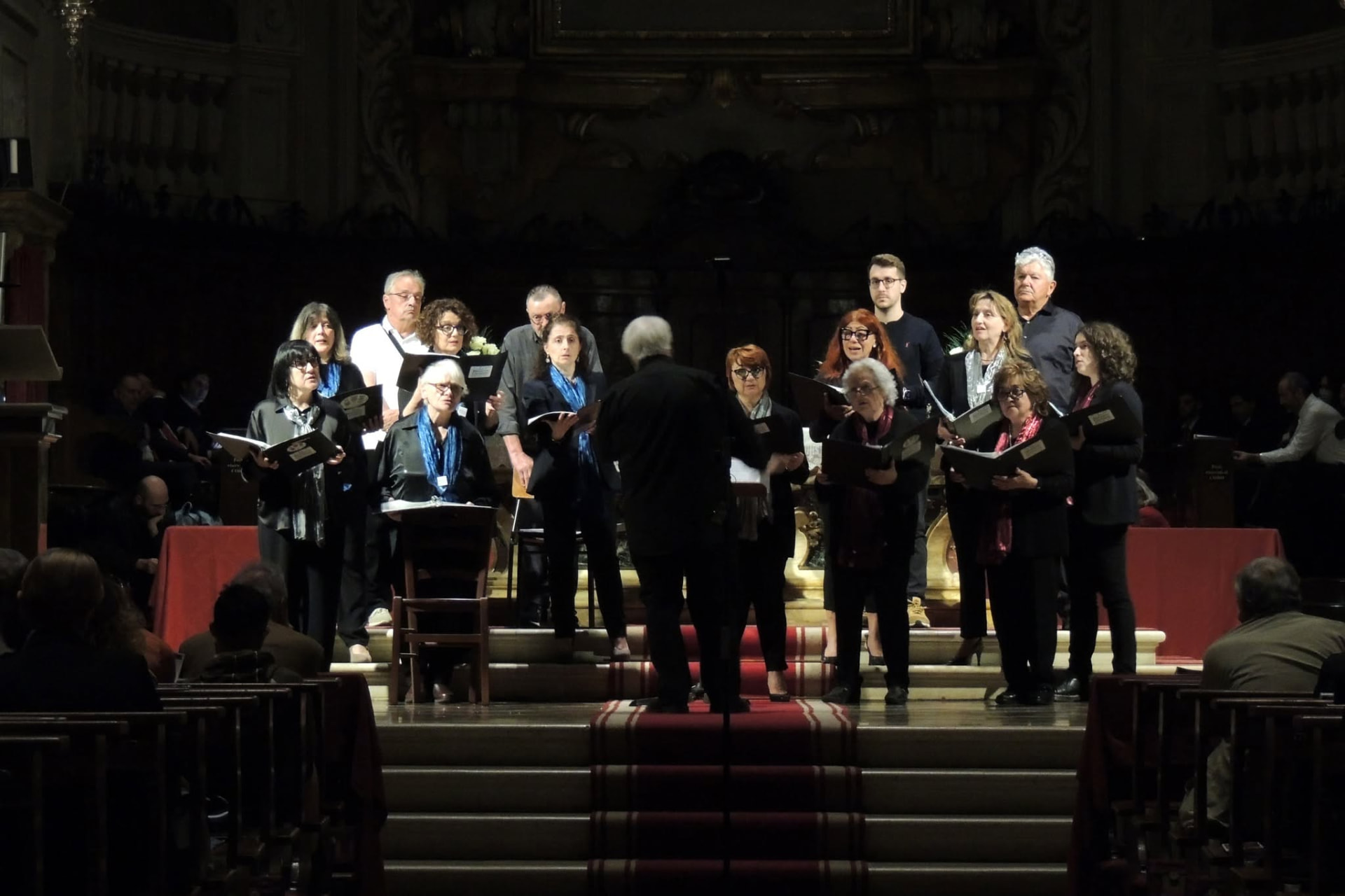Il maestro dirige il coro sui gradini dell'altare, tra le panche della chiesa